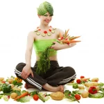 Junge Frau sitzt umgeben von Gemüse, hält dieses auf dem Arm und hat Kohlblätter auf dem Kopf.