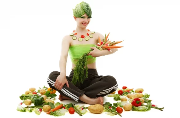 Young woman sits surrounded by vegetables, holds them in her arms and has cabbage leaves on her head.