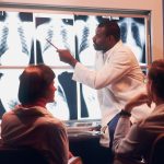 Arzt zeigt zwei sitzenden Kollegen etwas mit einem Stift auf einer erleuchteten Wand mit Röntgenbilder.