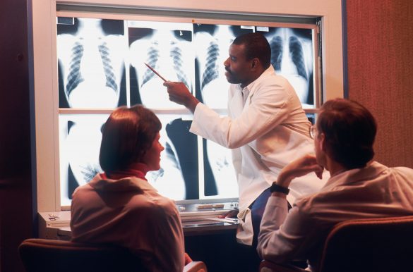 Arzt zeigt zwei sitzenden Kollegen etwas mit einem Stift auf einer erleuchteten Wand mit Röntgenbilder.